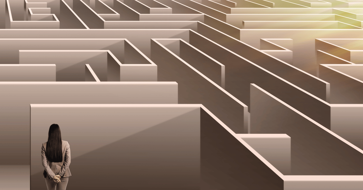 Woman in business attire standing before a large maze and considering which path to take.