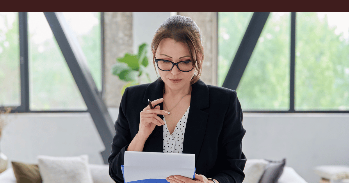 Woman wearing glasses reviewing notes on a tablet while thinking at a desk.